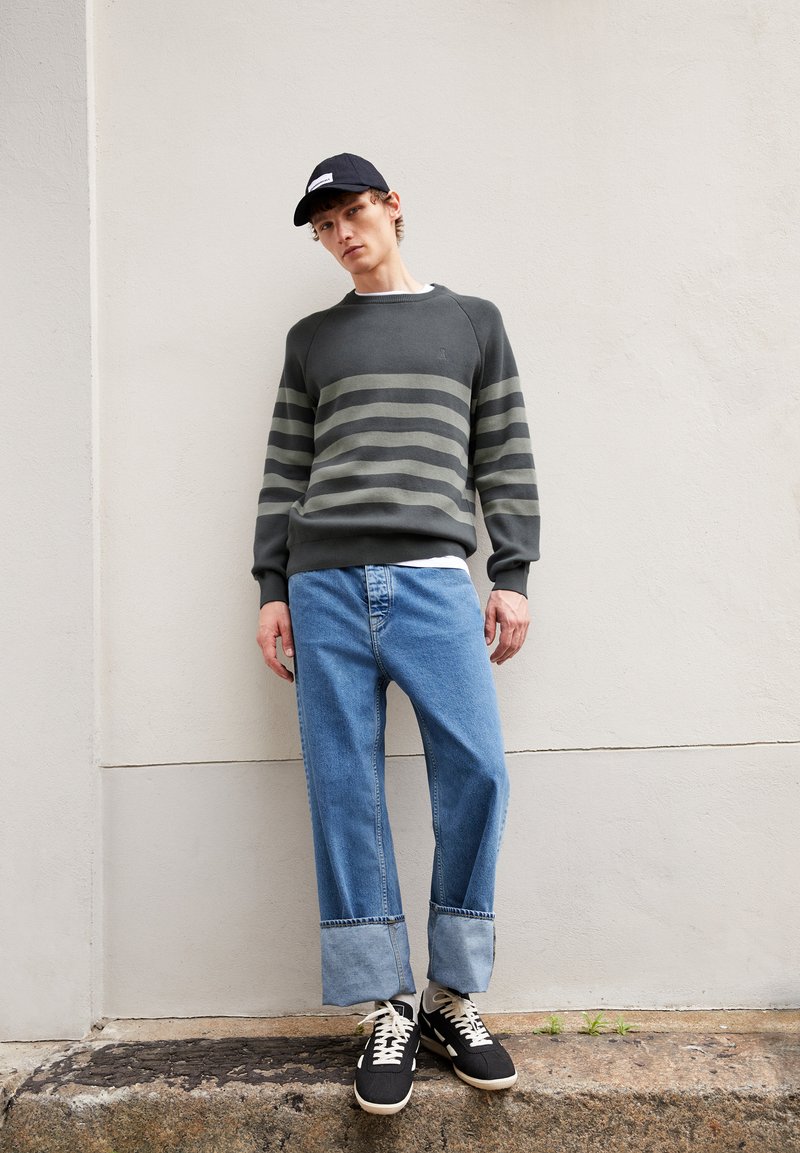 Grauer Pullover mit grünen Streifen, blaue gerade Jeans und schwarze Sneaker. Eine dunkle Kappe mit einem Logo rundet das Outfit ab.