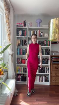 Eine Person in einem roten ärmellosen Kleid und Sneakers steht vor einem gefüllten weißen Bücherregal in einem sonnendurchfluteten Raum mit Pflanzen und einem kleinen Hund.
