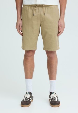 Moški, oblečen v bež kratke hlače z vrvico, bele nogavice in temnordeče superge, stoji ob enobarvnem svetlem ozadju.