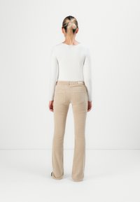 Woman with blonde hair in a bun wearing a white long-sleeve top and beige corduroy pants, standing facing a white wall.