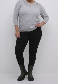 Pull gris avec des ornements floraux autour de l'encolure, associé à un pantalon noir ajusté et des bottines vert foncé montant à la cheville.