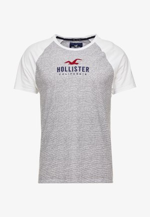 T-shirt raglan gris et blanc à manches courtes avec une mouette rouge et un texte bleu marine "Hollister California" sur la poitrine.