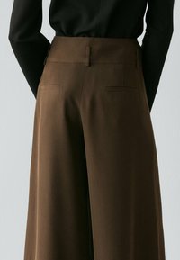 Vue de dos d'une personne portant un pantalon marron taille haute à jambes larges et un haut noir ajusté à manches longues sur un fond clair.