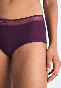 Lilla hipstertruser med teksturert stoff, med en blondelignende mesh-linning og en liten dekorativ ringdetalj. Glatt finish.