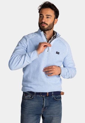 Lichtblauwe pullover met een ritskraag, gestructureerde stof en ribgebreide manchetten. Bevat een klein logopatch op de borst, gecombineerd met donkere spijkerbroek.