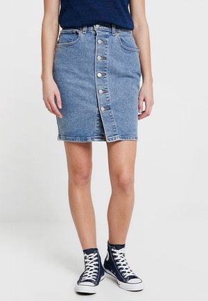 Kvinde iført en knaplukket denimnederdel, navyblå ærmeløs top og marineblå høje sneakers med hvide snørebånd, stående mod en ensfarvet baggrund.