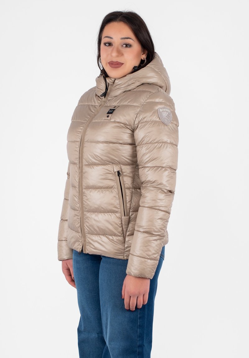 Giacca a vento imbottita beige con cappuccio, design trapuntato, tasche con zip frontali e patch con logo sulla spalla. Realizzata in materiale lucido e isolante.