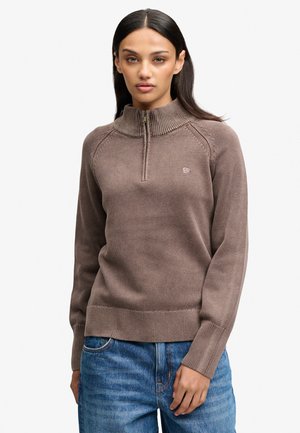 Kvinde iført en brun striktrøje med halvlange lynlås og blå jeans, stående mod en ensfarvet hvid baggrund.