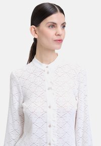 Blusa bianca in pizzo con colletto alla mandarina, bottoni perla e delicati motivi floreali, dotata di maniche lunghe e una finitura testurizzata.
