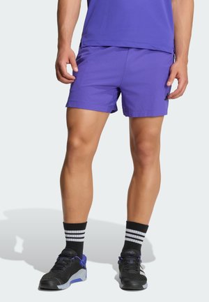 Mann trägt lila Sportshorts, passendes Shirt, schwarz-weiß gestreifte Socken und schwarze sowie lila Sportschuhe, steht in entspannter Haltung.