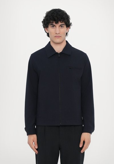 Navy zip-up jacket in soft fabric, featuring a pointed collar, front pocket with zipper, and long sleeves. Minimalist design, relaxed fit.