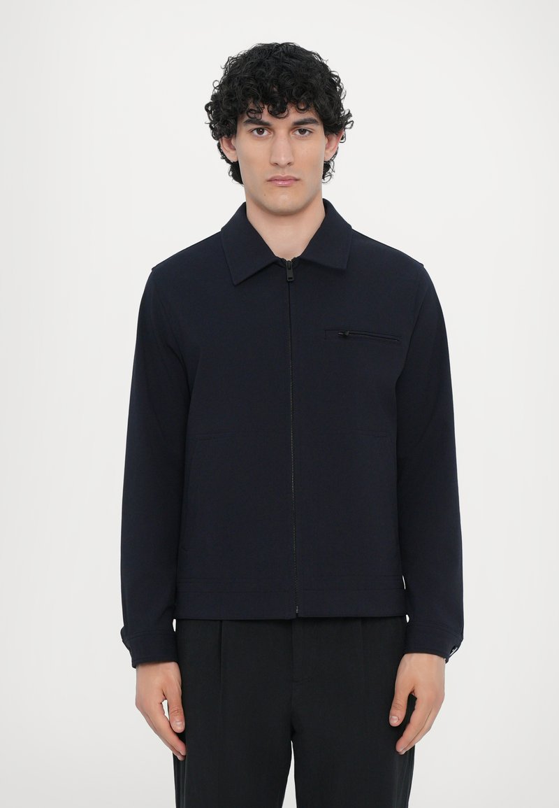 Modra zip-jakna iz mehkega materiala, z ostrim ovratnikom, sprednjim žepom z zadrgo in dolgimi rokavi. Minimalističen dizajn, sproščen kroj.