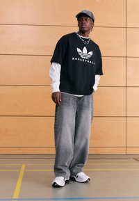 Svart t-shirt med kort ärm och vit text "BASKETBALL" samt logotyp, lager-på-lager över en vit långärmad tröja, kombinerad med ljusgråa baggy-jeans och sneakers.