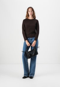 Brun en tettsittende genser, høy midje blå jeans og en teksturert svart håndveske med snøring. Modellen står mot en hvit bakgrunn.