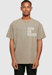 Overdimensioneret beige t-shirt lavet af bomuld, med rund hals og korte ærmer. Teksten "NOW OR NEVER" trykt i hvidt på forsiden.