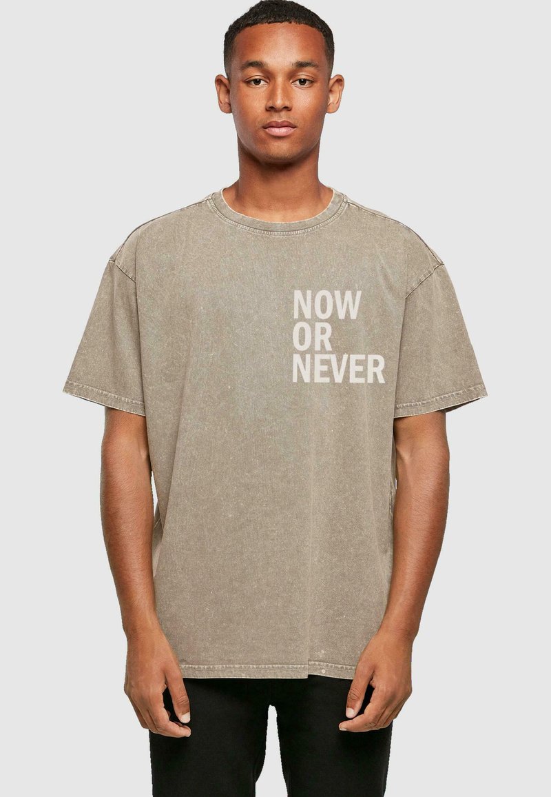 Overdimensioneret beige t-shirt lavet af bomuld, med rund hals og korte ærmer. Teksten "NOW OR NEVER" trykt i hvidt på forsiden.