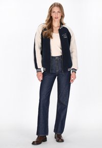 Marineblå og creme fleecejakke med lynlås foran, stribede manchetter og broderede detaljer, parret med højtaljede jeans og brune støvler.