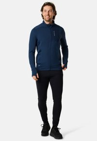 Dunkelblaue Zip-Jacke mit hohem Kragen, langen Ärmeln und Fronttasche, kombiniert mit schwarzer, enganliegender Leggings und schwarzen Sportschuhen.