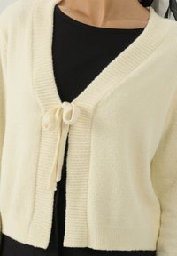 Cardigan color crema con texture a coste e chiusura a cravatta nella parte anteriore. Presenta un collo a V profondo e un tessuto morbido e soffice.