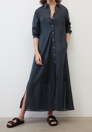 Długa, ciemnoniebieska sukienka z guzikami, wykonana z lekkiego materiału, z kołnierzykiem, długimi rękawami, bokami z rozcięciami i luźnym krojem. Na nogach czarne sandały.
