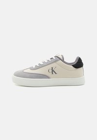 Tenisky Calvin Klein s béžovou plátnovou svrchní částí a šedou semišovou patou, kulatou špičkou, bílými tkaničkami, černými akcenty na patě a logem na boku.