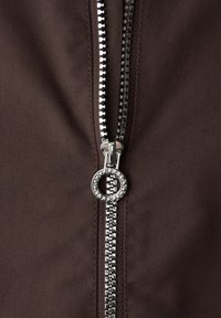 Nærbillede af sølvmetal lynlås delvist åbnet på mørkebrunt stof med en rund trækketag, der har indgraveret tekst.