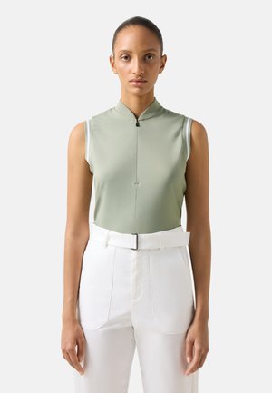 Vrouw draagt een mouwloze lichtgroene top met rits aan de voorkant en witte hoge taillebroek met riem, staand tegen een effen achtergrond.