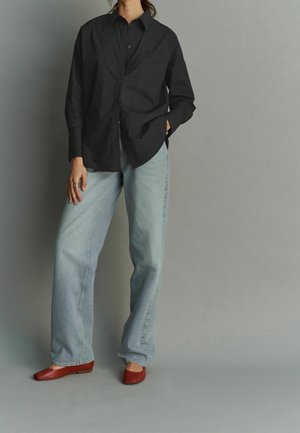 Personne portant une chemise noire ample à boutons, un jean bleu clair à jambes larges, et des chaussures plates rouges, debout devant un fond gris.