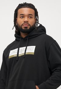 Black hoodie made of cotton with a front pocket, featuring a white and gold logo patch reading "JACK & JONES" for branding.
