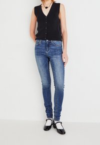 Top negro sin mangas con escote en V y cierre de botones, combinado con jeans de mezclilla azul ajustados. El modelo lleva zapatos negros con calcetines blancos.