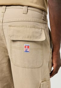 Beige cargobyxor med en stor bakficka och en Wrangler-logotyp patch. Tillverkade av texturerat tyg, med förstärkta sömmar och justerbar midja.