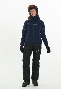 Marinblå skidjacka med kontrasterande ljusblå sömmar, svarta skidbyxor, svarta handskar och en svart hjälm med bruna goggles. Vinter sportkläder.