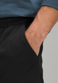 Shorts de sport noirs avec une texture lisse, dotés de poches latérales et d'une taille élastique. Main reposant dans la poche.
