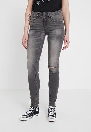 Jeans ajustados grises desgastados con rasgadura en la rodilla, diseño de cinco bolsillos y trabillas para el cinturón. Combinados con zapatillas altas negras.