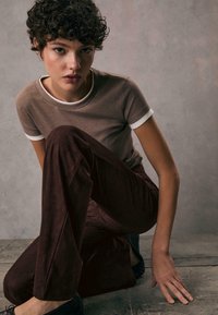 Knielanger, taillierter brauner Ribbel-Top mit weißen Akzenten, kombiniert mit hochtaillierten, dunkelbraunen Samenhosen. Das Model kniet auf einer strukturierten Oberfläche.