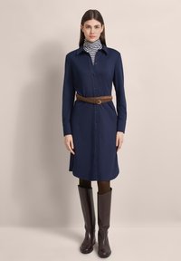 Hemdblusenkleid in Marineblau mit langen Ärmeln, einem braunen Gürtel in der Taille und braunen knielangen Stiefeln, kombiniert mit einem schwarz-weiß gestreiften Rollkragenpullover.