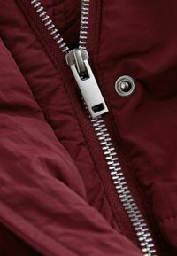 Das burgunderrote, gesteppte Gewebe verfügt über einen silbernen Metallreißverschluss mit rechteckigem Zuganhänger und eine Druckknopfdetaillierung an der Seite.