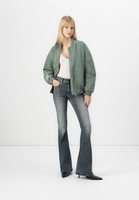 Giacca bomber verde con zip frontale, canotta grigia, jeans in denim scuro a gamba svasata e tacchi neri a punta. Tessuti lisci, vestibilità rilassata.