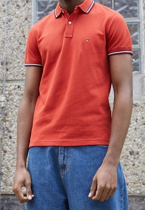 Rotes Poloshirt mit klassischem Kragen, kurzen Ärmeln und marineblauen sowie weißen Streifenakzenten. Getragen mit blauen Jeans aus Denim.