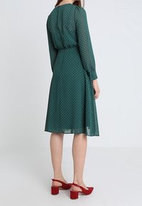 Robe verte à pois avec des manches longues, taille cintrée, tissu fluide et jupe à mi-genoux. Assortie à des sandales à talons carrés rouges.