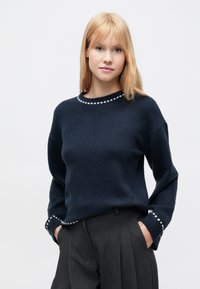 Pull bleu marine avec une finition texturée, encolure ronde, et surpiqûres blanches en accent à l'ourlet et aux poignets, associé à un pantalon gris.
