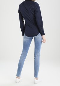 Granatowa koszula z długim rękawem, dopasowany krój, z mankietami na guziki; noszona z jasnoniebieskimi skinny jeansami i białymi trampkami. Widok od tyłu.