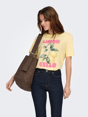 Mujer con camiseta gráfica amarilla con diseño de limón y jeans oscuros, llevando un gran bolso marrón al hombro, mirando hacia abajo, fondo claro.