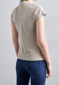 Beige blouse met korte mouwen en een getailleerd ontwerp, gladde textuur en hoge halslijn, gecombineerd met donkerblauwe slim-fit jeans.