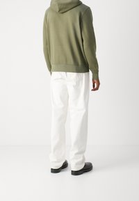 Felpa verde oliva con tasca a canguro, indossata con pantaloni bianchi larghi e scarpe nere; dettagli di cucitura visibili e cappuccio.