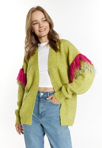 Limegrüner gestrickter Cardigan mit übergroßen Ärmeln, der pinke und graue Fransenakzente aufweist; getragen über einem weißen Oberteil und kombiniert mit hellblauen Jeans.