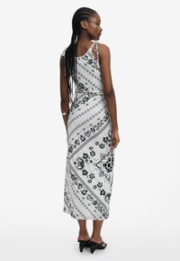 Maxikleid in Weiß mit schwarzen Blumen- und geometrischen Mustern, ausgestattet mit dünnen Trägern und einer glatten Stofftextur, kombiniert mit schwarzen High-Heelsandalen.