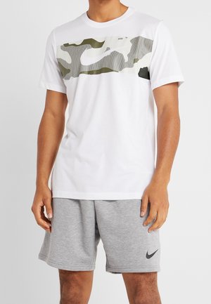 Mężczyzna ubrany w białą koszulkę z szaro-zielonym paskiem w kamuflażu oraz szare szorty z czarnymi logo Nike, stojący na jednolitym tle.