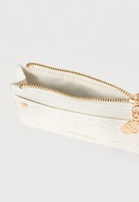 Pochette blanche zippée avec un motif diamant texturé, une quincaillerie dorée et un charm's en accent. L'intérieur est doté d'une doublure lisse.