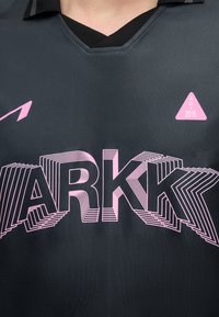 Μαύρο πουκάμισο με ροζ λογότυπο ARKK σε στιβαρό κείμενο και ένα μικρό ροζ τρίγωνο με "AC 2015" στο στήθος, φορεμένο από ένα άτομο.